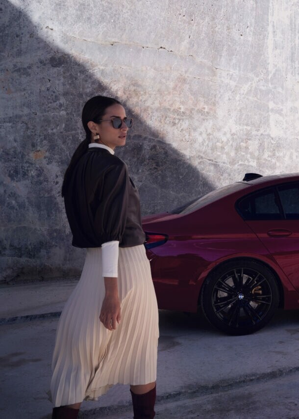 Eine elegante Frau mit Sonnenbrille vor einem roten BMW vor einer Betonwand.