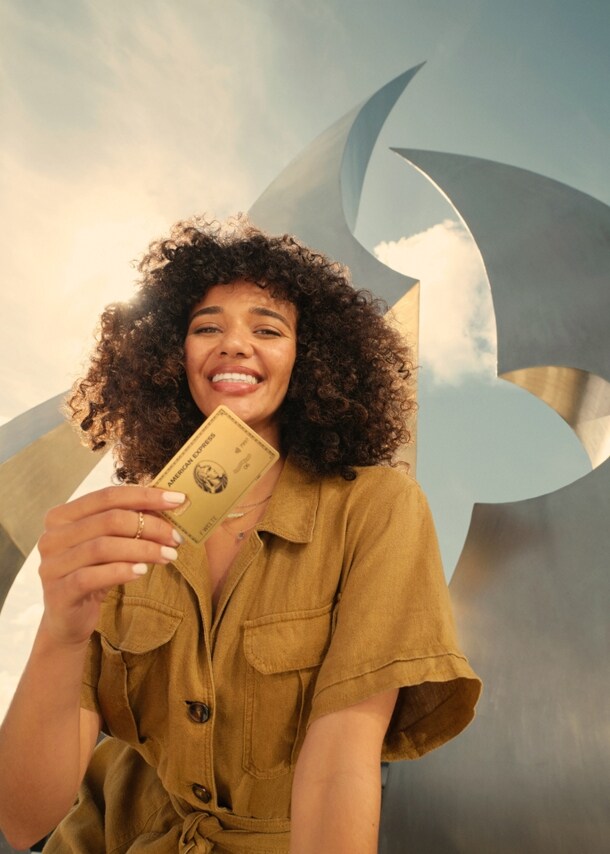 Eine lächelnde junge Frau hält eine goldene Kreditkarte von American Express in der Hand, im Hintergrund eine moderne Skulptur im Freien bei Sonnenschein.