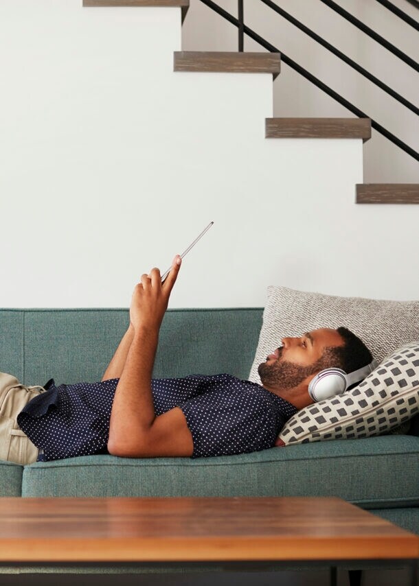 Ein Mann liegt mit Kopfhörern über den Ohren auf einem Sofa und liest.