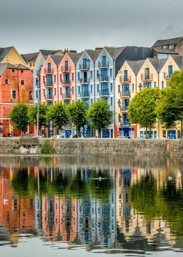Bunte Wohngebäude in verschiedenen Farben spiegeln sich im ruhigen Wasser eines Hafens. Bäume stehen entlang der Uferpromenade.
