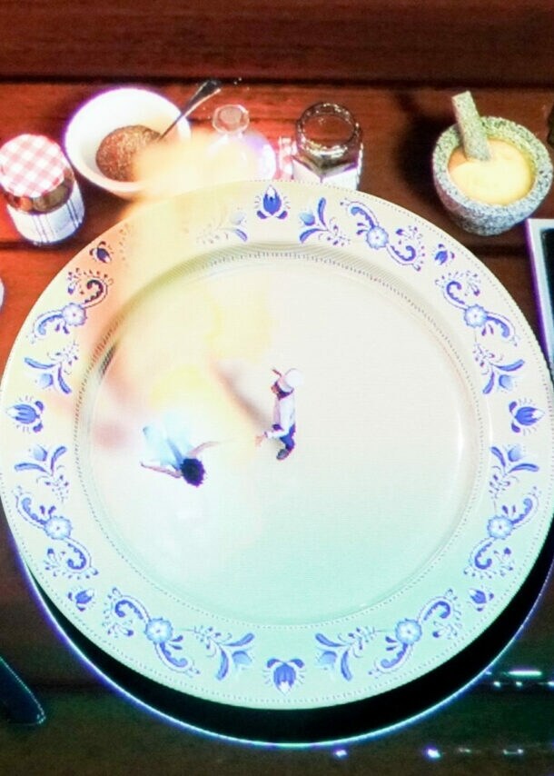 Ein gedeckter Tisch mit einem großen weißen Teller, auf dem zwei kleine Figuren stehen. Daneben befinden sich ein Messer, eine Gabel, ein Glas, Gewürzgläser und eine Pfanne mit einem Ei.