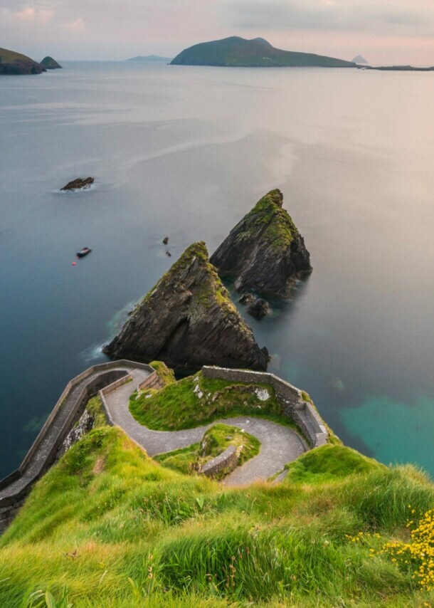 Blick auf eine Küstenlandschaft mit grünem Hügel, steilen Klippen und ruhigem Wasser. Im Hintergrund sind mehrere Inseln und ein Boot sichtbar.