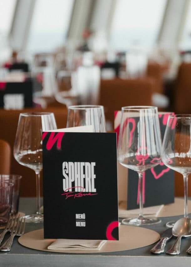 Tischgedeck in einem Restaurant mit Gläsern, einer Flasche und einer Speisekarte mit dem Titel 'SPHERE'. Im Hintergrund sind weitere Tische sichtbar.