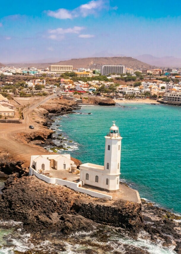 Luftaufnahme eines Leuchtturms auf einer felsigen Küste, umgeben von türkisblauem Wasser und Wellen, mit einer Stadt im Hintergrund.