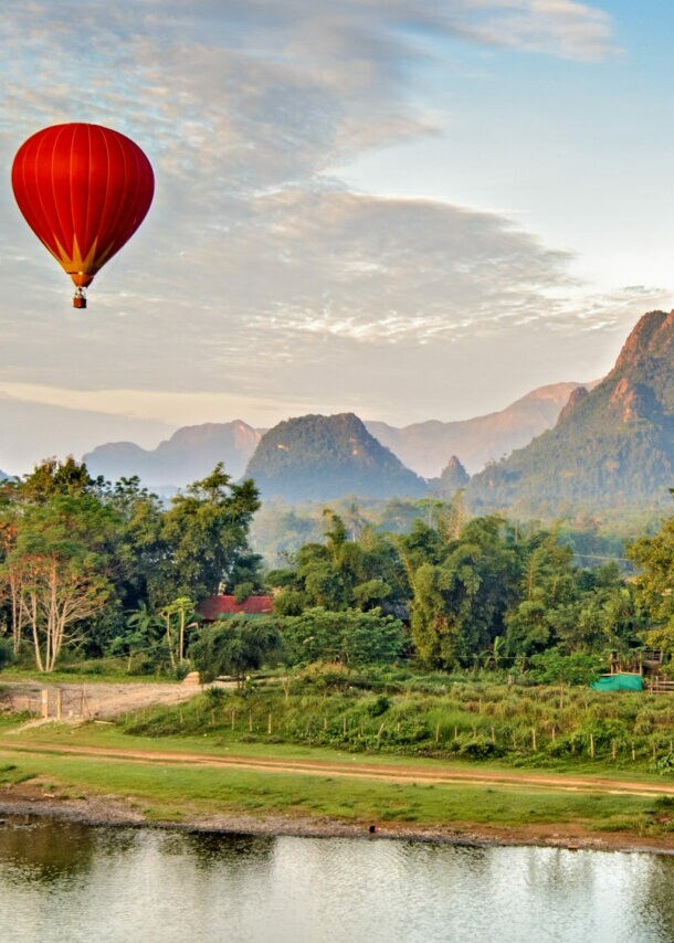 Ein roter Heißluftballon schwebt über einer grünen Landschaft mit Bergen und einem ruhigen Fluss.