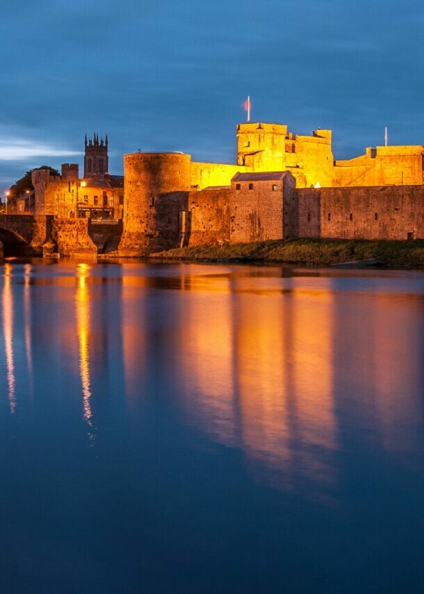 Blick auf eine beleuchtete Burg am Wasser bei Dämmerung. Die Burg hat Türme und eine Brücke im Vordergrund. Reflektionen im Wasser sind sichtbar.