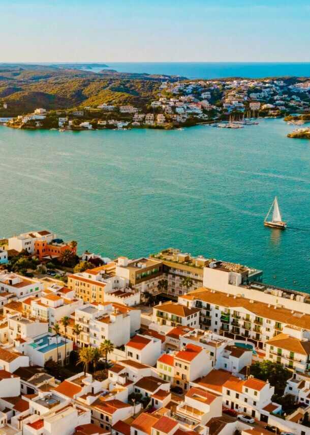 Luftaufnahme einer Küstenstadt mit weißen Gebäuden und roten Dächern, umgeben von Wasser und Hügeln. Ein Segelboot ist auf dem Wasser sichtbar.