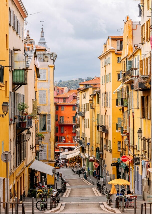 Blick auf eine enge, gepflasterte Straße mit bunten Gebäuden in Gelb- und Orangetönen. Auf beiden Seiten stehen Balkone mit Pflanzen. Im Hintergrund sind Tische und Stühle eines Cafés zu sehen.