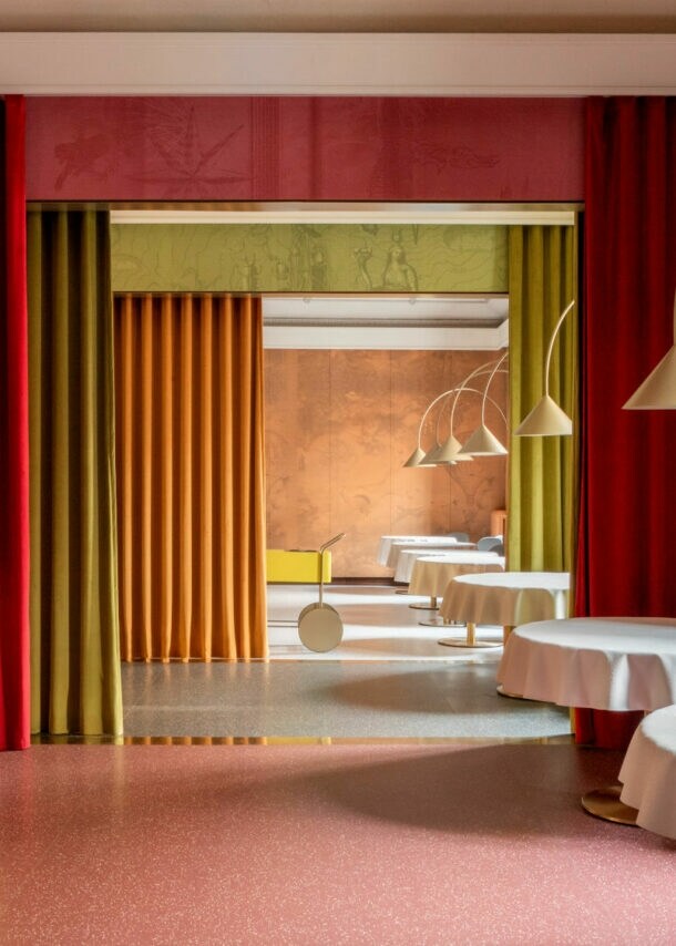 Ein eleganter Innenraum mit runden Tischen, die mit weißen Tischdecken bedeckt sind. An den Wänden hängen rote und grüne Vorhänge. Im Hintergrund sind gelbe und orangefarbene Wände sichtbar.