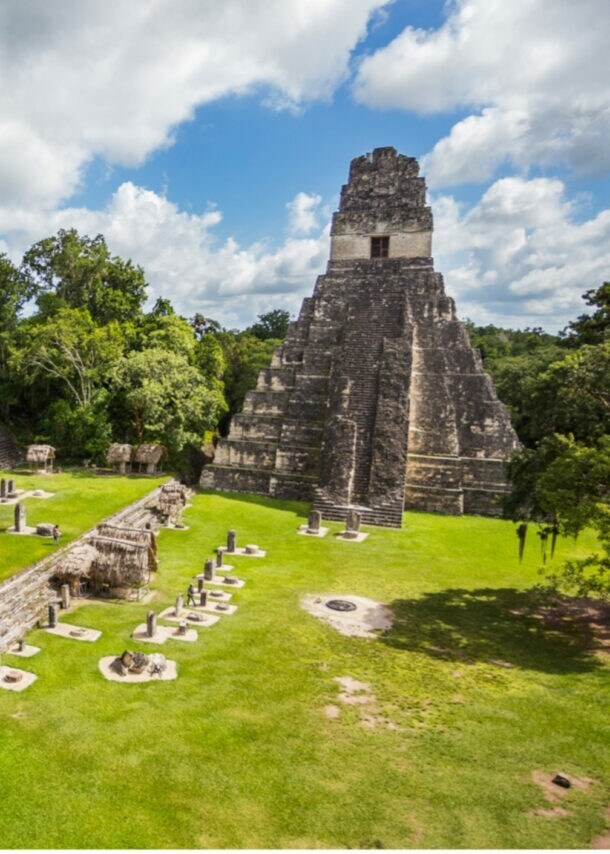 Maya-Tempelanlage mit Stufenpyramide und mehreren Steinstrukturen umgeben von grünem Gras und Bäumen unter blauem Himmel mit Wolken
