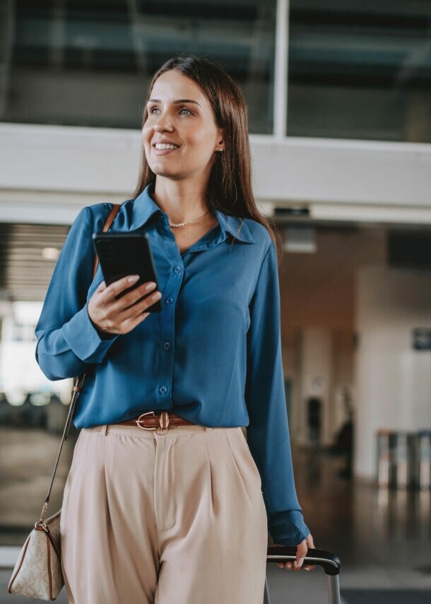 Eine elegant gekleidete Frau mit Smartphone steht vor dem Eingang der Ankunftshalle eines Flughafen-Terminals.