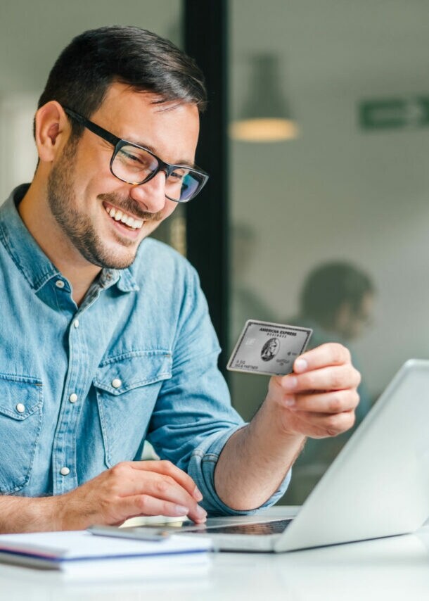 Ein gut gelaunter Mann im Jeanshemd sitzt an einem Tisch vor einem Laptop in einem Büro und hält die Business Platinum Card von American Express in der Hand.