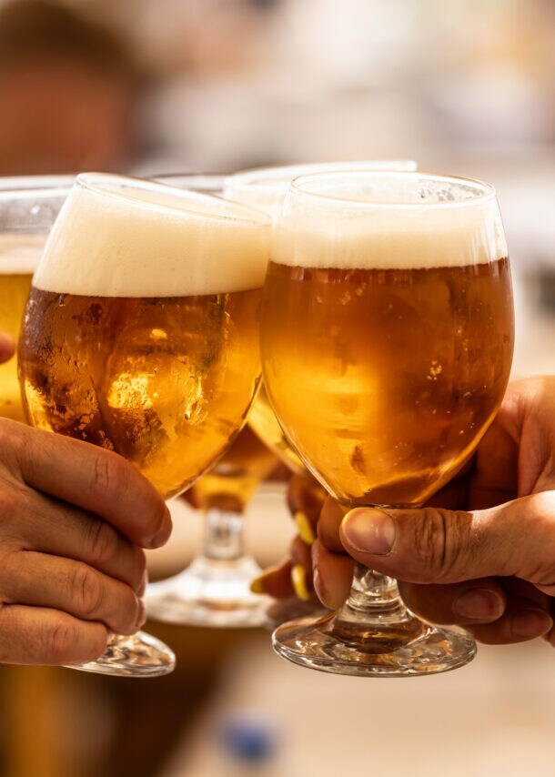 Vier Gläser mit Bier werden zum Anstoßen gehalten. Zwei Gläser sind in der Hand einer Person mit einem blauen Armband. Die Biere haben eine goldene Farbe und eine schaumige Oberseite.