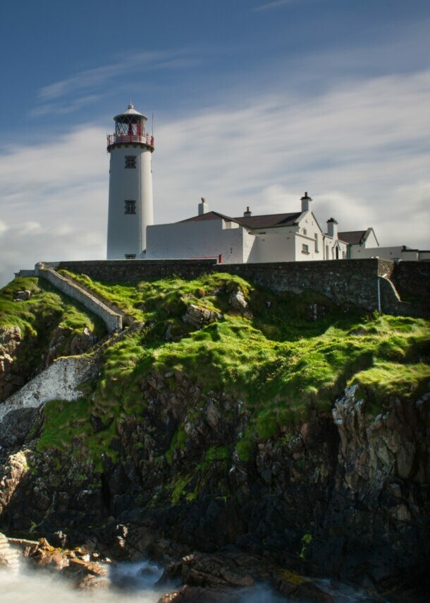 Ein Leuchtturm mit rotem Licht auf einem Hügel, umgeben von grünem Gras und steiniger Küste, mit Wellen, die gegen die Felsen schlagen.