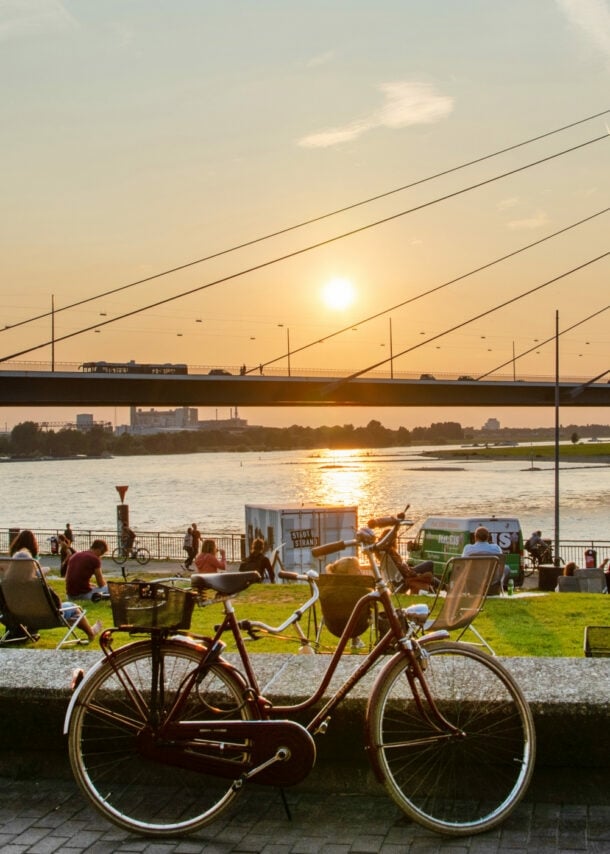 Ein rotes Fahrrad steht auf dem Bürgersteig, im Hintergrund ist eine Brücke und der Sonnenuntergang über dem Wasser zu sehen. Menschen entspannen auf der Wiese.