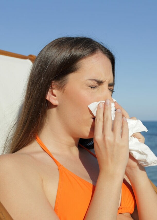 Eine Frau in einem orangefarbenen Bikini sitzt auf einer Liege am Strand und hält ein Papiertaschentuch in der Hand. Im Hintergrund sind Felsen und das Meer sichtbar.