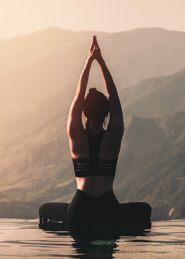 Eine Person in schwarzer Sportbekleidung sitzt im Lotussitz am Rand eines Infinity-Pools. Sie hat die Hände über dem Kopf zusammengelegt und blickt auf die Berge im Hintergrund.
