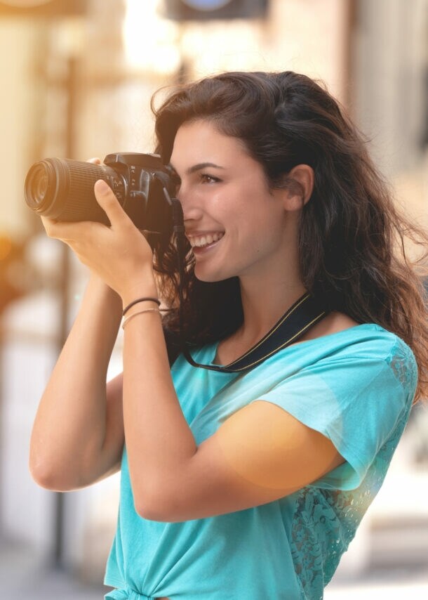 In einer urbanen Umgebung hält eine Frau in einem mintfarbenen T-Shirt eine Kamera vors Gesicht und blickt durch die Linse.