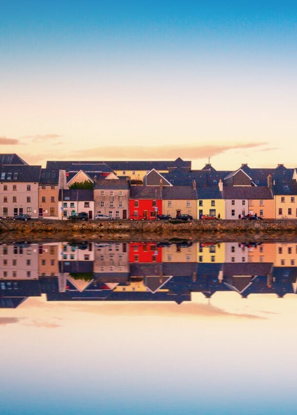 Bunte Häuser in Galway, Irland, entlang eines ruhigen Wassers mit Spiegelung. Die Gebäude sind in verschiedenen Farben, darunter Rot, Gelb und Weiß.