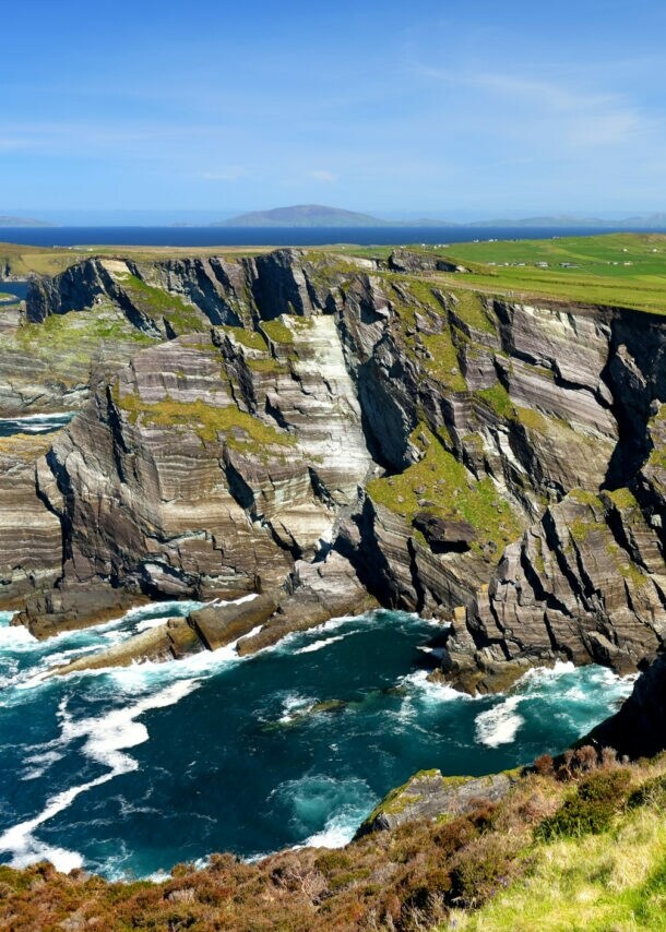 Luftaufnahme einer Küstenlandschaft mit steilen Klippen, grünem Gras und einem schmalen Weg, der entlang der Klippen verläuft. Im Hintergrund sind sanfte Hügel und ein blauer Himmel zu sehen.