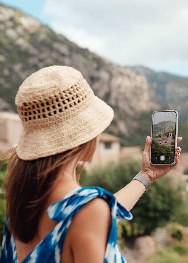 Frau mit Strohhut und blauem Kleid fotografiert Bergdorf mit Smartphone