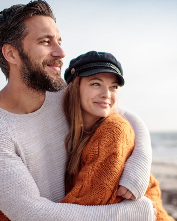 Ein sich umarmendes Paar in Pullovern steht glücklich lächelnd an einem Sandstrand und blickt aufs Meer.