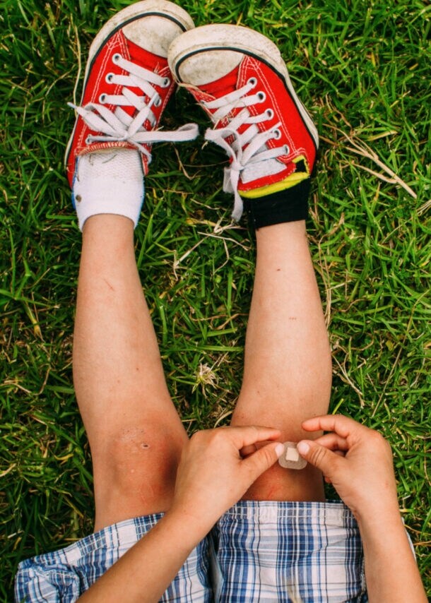 Kind sitzt auf Gras, trägt rot-weiße Turnschuhe und blau karierten Rock, hält Pflaster an Knie.