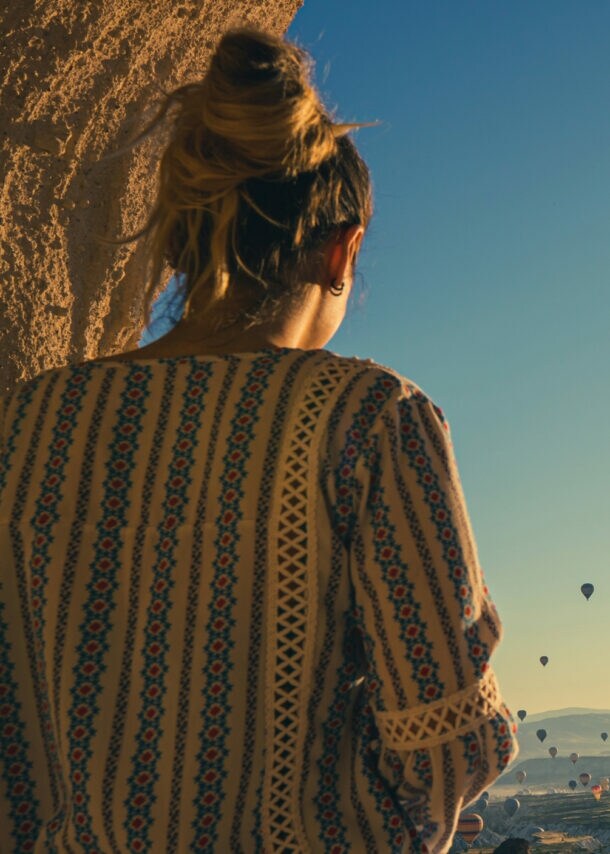 Eine Person mit einem langen, gemusterten Oberteil steht mit dem Rücken zur Kamera und blickt auf eine Landschaft mit vielen Heißluftballons im Himmel bei Sonnenaufgang.