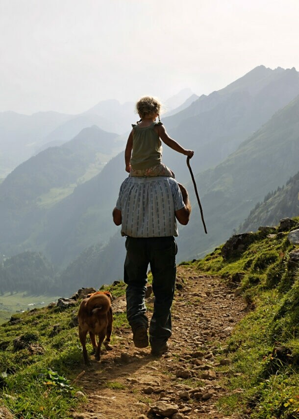 Ein Erwachsener trägt ein Kind auf den Schultern, während sie einen Wanderweg entlanggehen. Ein Hund läuft neben ihnen. Die Landschaft zeigt Berge und Täler im Hintergrund.