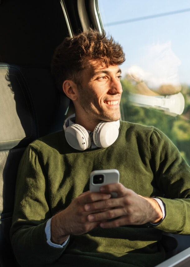 Junger Mann mit grünem Pullover und weißen Kopfhörern sitzt im Zug mit Smartphone in der Hand und blickt aus dem Fenster.