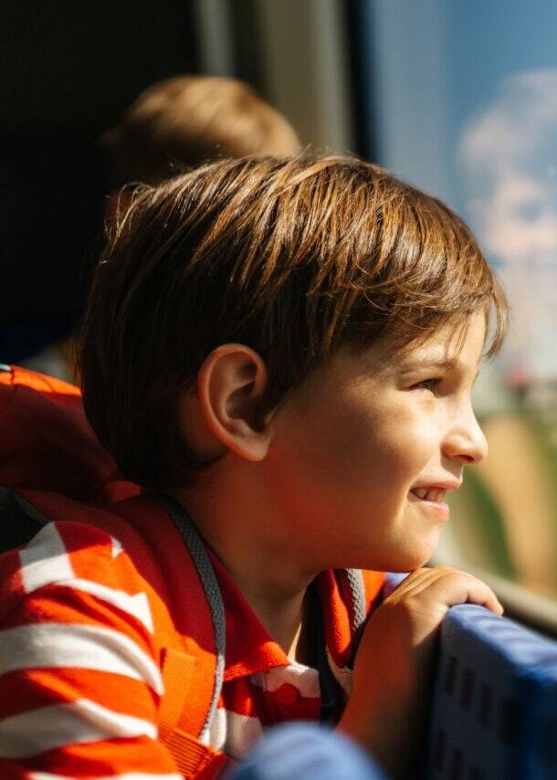 Ein Kind mit gestreiftem Oberteil lehnt sich am Zugfenster und schaut nach draußen. Im Hintergrund ist ein weiteres Kind zu sehen.