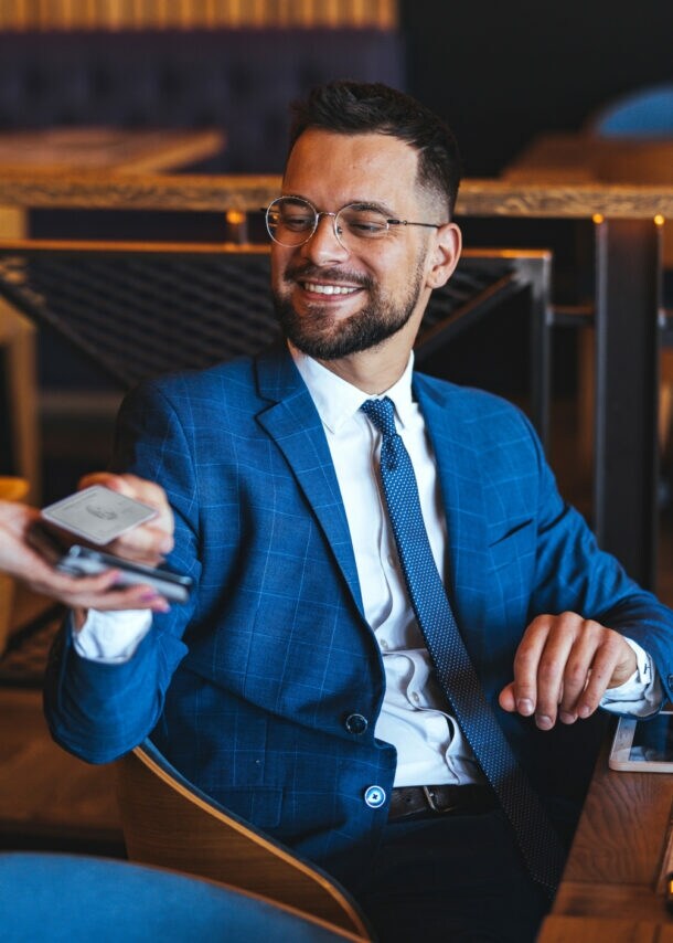 Ein Mann im blauen Anzug sitzt an einem Tisch in einem Bistro und hält eine Kreditkarte an ein Kartenlesegerät, das eine Person ihm hinhält.