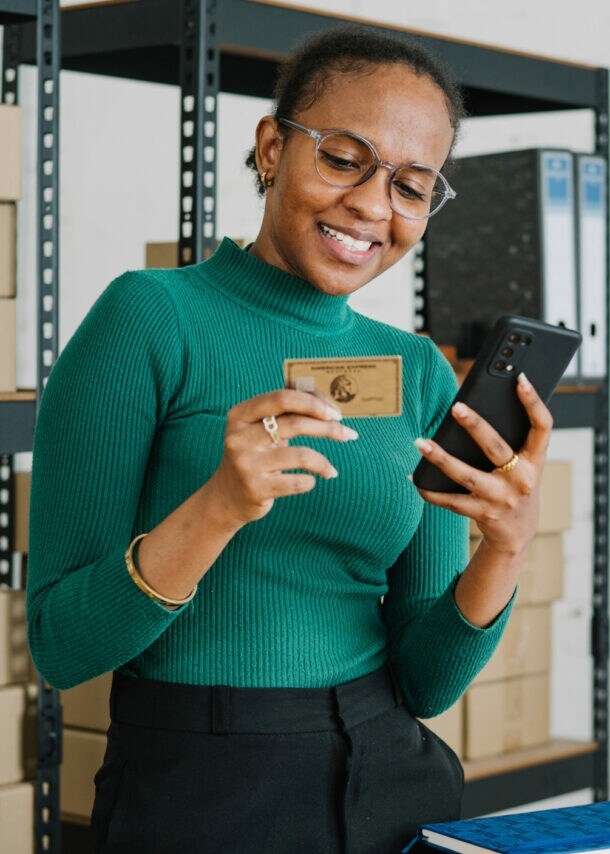 Eine lächelnde Frau im grünen Shirt steht vor einem Lagerregal mit Kartons und hält ein Smartphone sowie eine goldene Kreditkarte von American Express vor sich in den Händen.