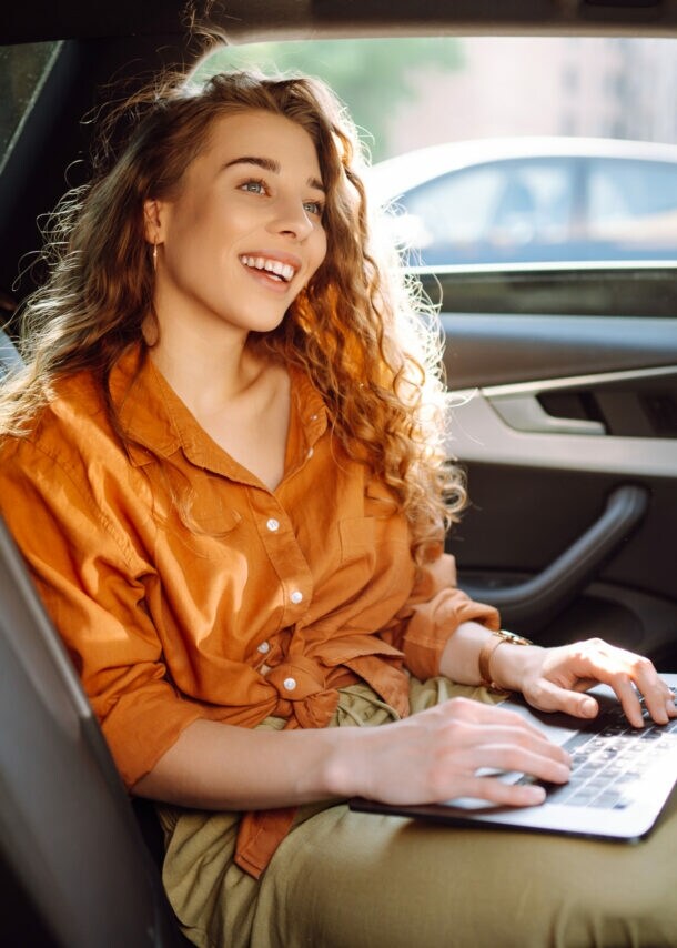 Eine lachende Frau in orangefarbener Bluse sitzt auf dem Rücksitz eines Autos mit einem aufgeklappten Laptop auf ihrem Schoß.