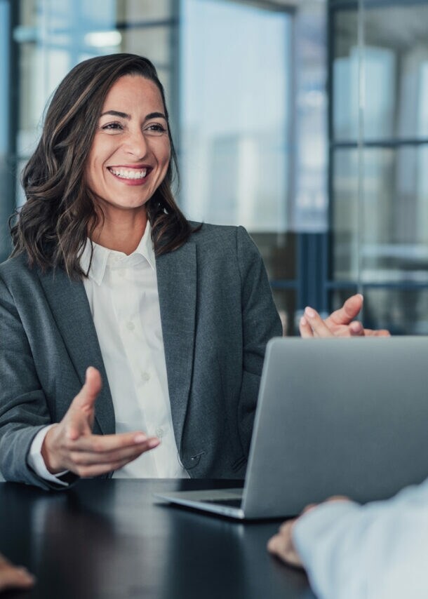 Eine lachende Frau im grauen Blazer sitzt vor einem Laptop an einem Tisch gegenüber zwei Personen in einem Raum mit Glaswänden.