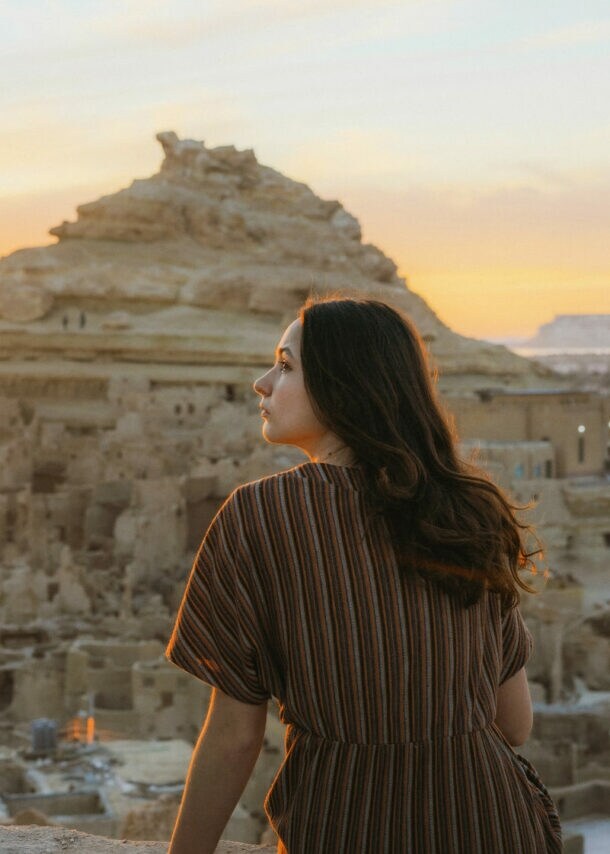 Frau mit braunem gestreiftem Kleid sitzt auf einer Mauer und blickt auf die Felsenstadt in der Siwa-Oase bei Sonnenuntergang in Ägypten.