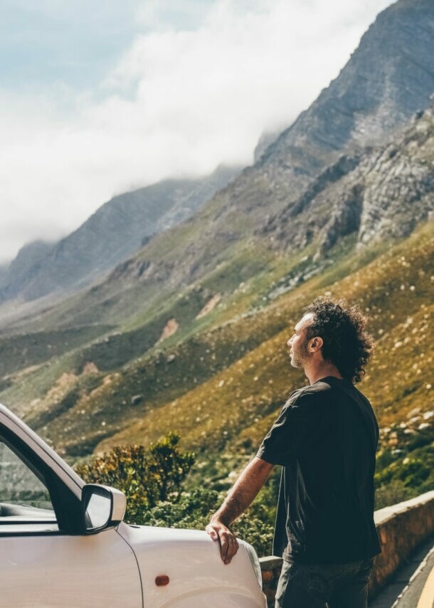 Mann lehnt an weißem Auto an einer Bergstraße, umgeben von steilen Felsen und bewölktem Himmel.