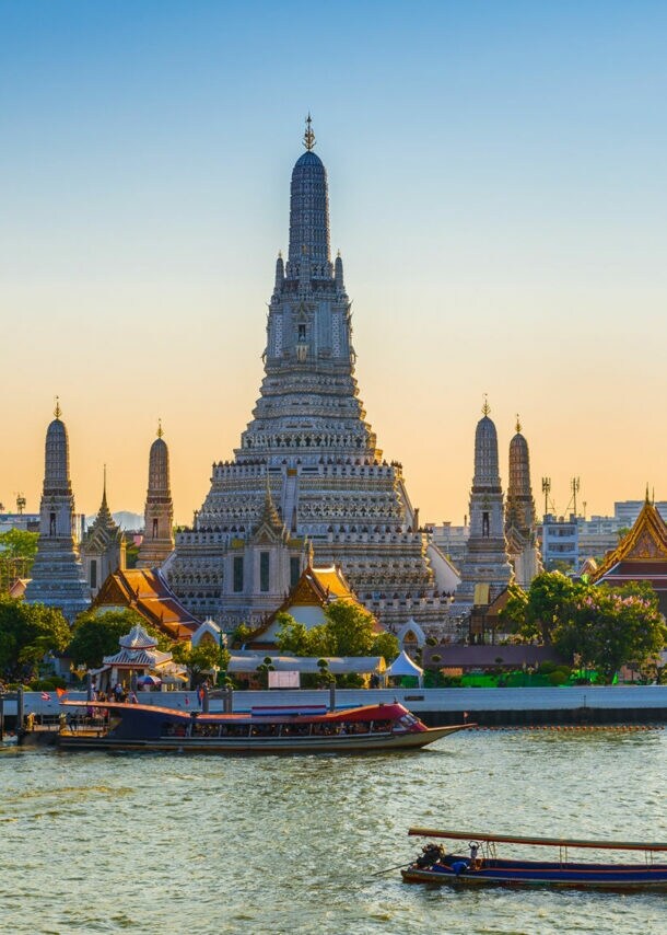 Vor einem großen Fluss mit Booten ragt ein großer Tempel empor, er ist umgeben von kleineren Türmen und Häusern mit spitzen Dächern.