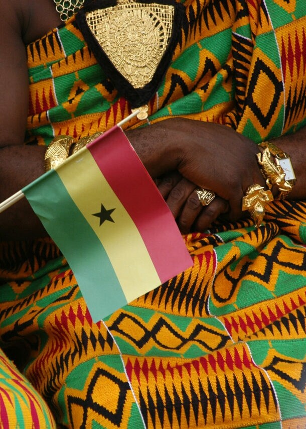 Oberkörper zweier sitzender Frauen in traditionellen ghanaischen Kleidern aus buntem Kente-Stoff und Goldschmuck, davor eine kleine ghanaische Flagge aus Papier in einer Hand gehalten.