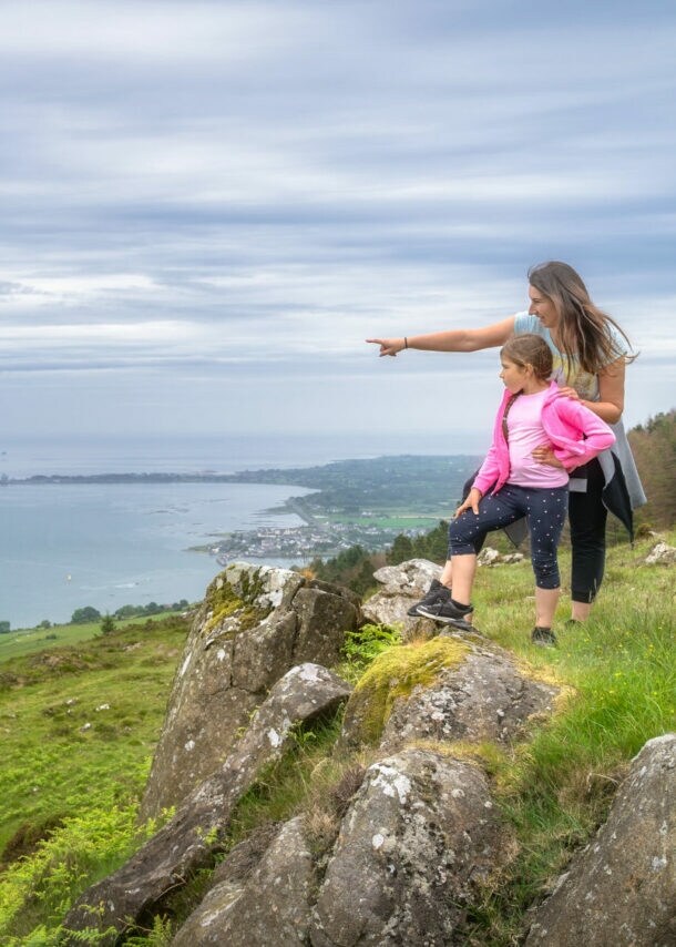 Eine Frau steht mit einem Mädchen auf einem Hügel in einer grünen Graslandschaft an einer Küste und zeigt hinaus aufs Meer.
