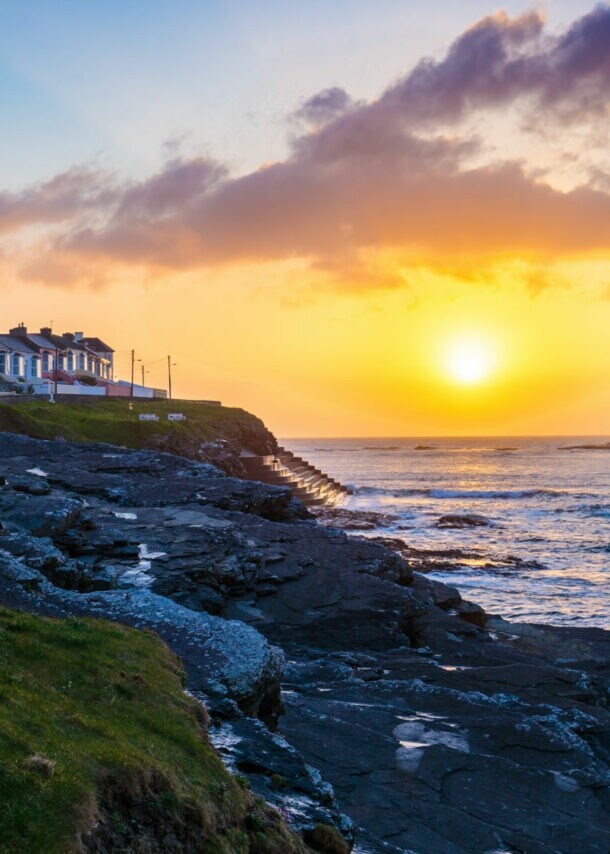 Sonnenuntergang über der Küste von Kilkee mit Reihenhäusern auf einer Steinmauer und Felsen am Meer