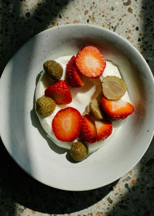 Schale mit Joghurt, belegt mit geschnittenen Erdbeeren und kleinen grünen Früchten, auf marmoriertem Untergrund mit Schatten.