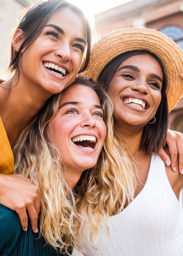 Drei lachende junge Frauen stehen sich umarmend auf einem Platz.
