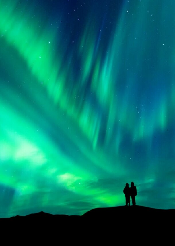 Zwei Personen stehen auf einem Hügel und beobachten das grün leuchtende Nordlicht am Nachthimmel in Norwegen.