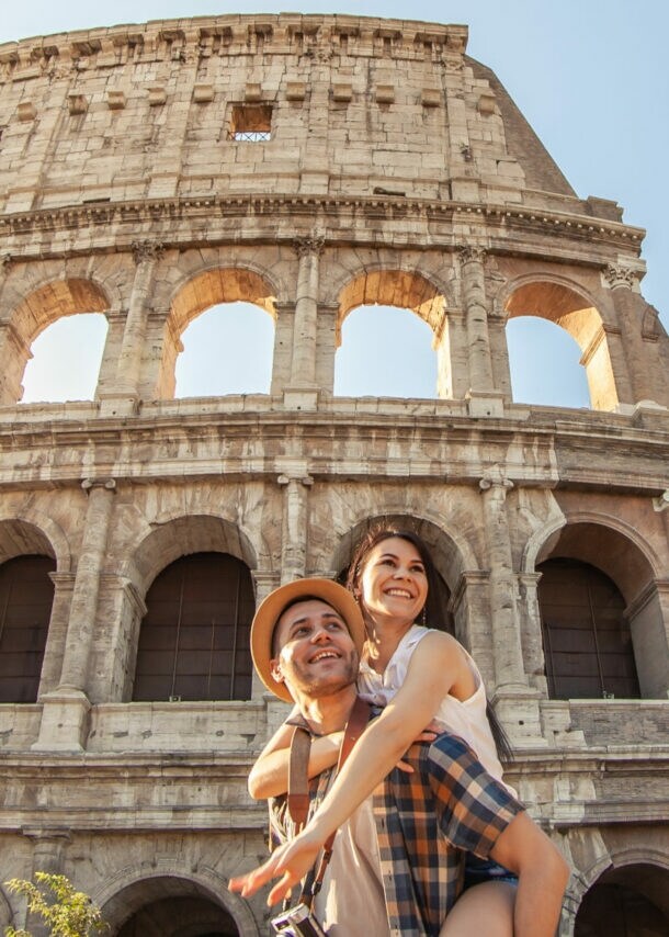 Das Kolosseum in Rom bei Sonnenlicht, davor ein Mann mit kariertem Hemd, der eine Person auf dem Rücken trägt.