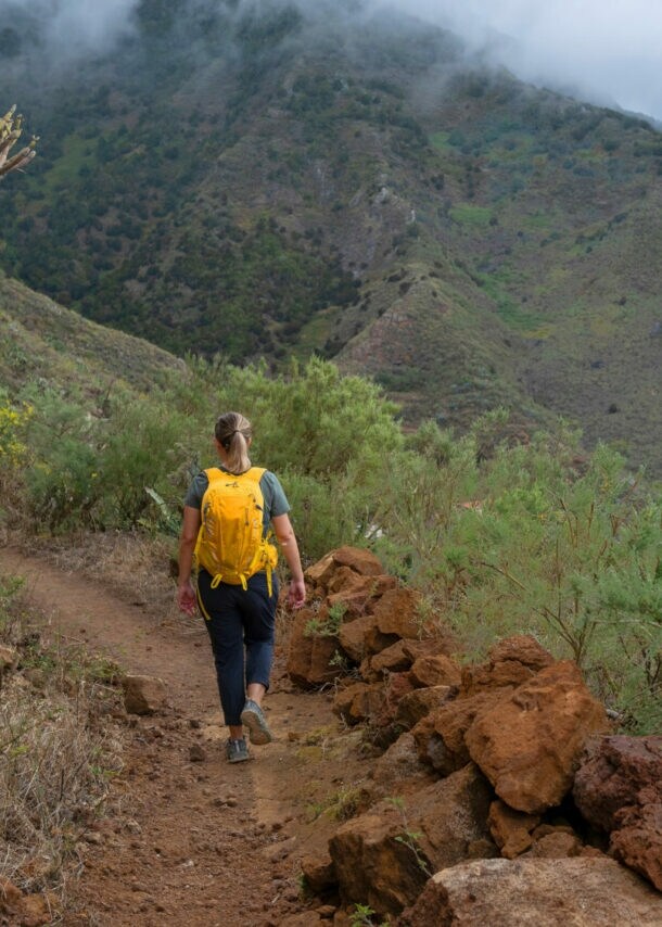 Eine Person mit gelbem Rucksack geht auf einem Pfad in bergiger Landschaft auf Teneriffa. Der Himmer ist bewölkt, am Wegesrand ist grüne Vegetation.