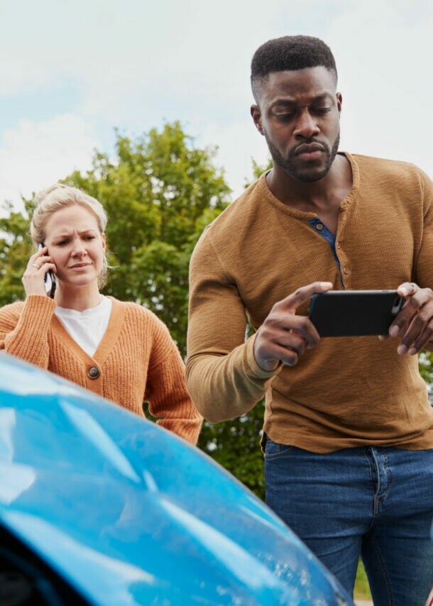 Ein Mann und einer Frau mit Smartphones stehen hinter zwei Autos mit Blechschäden, er macht ein Foto vom Schaden.