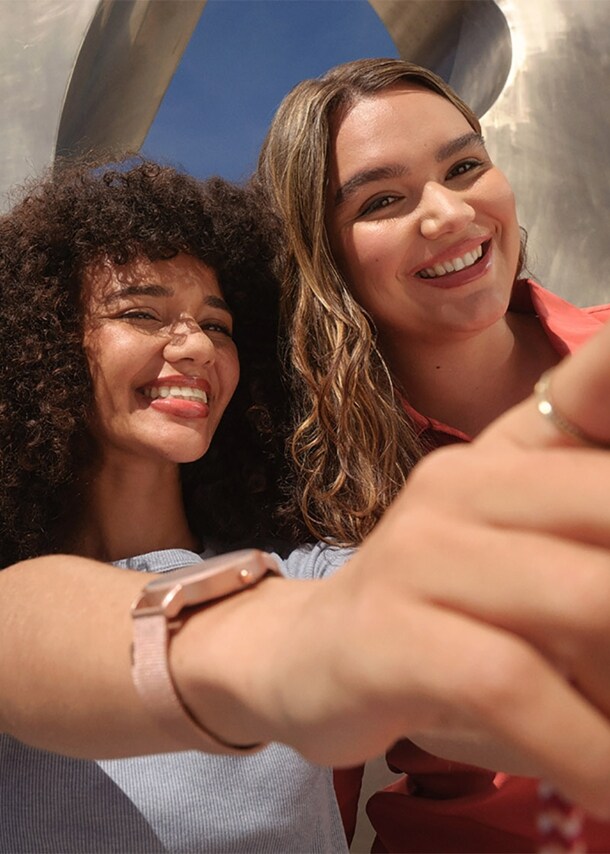 Zwei junge Frauen machen ein Selfie mir einem rosafarbenen Smartphone vor einer Metallskulptur unter freiem Himmel.