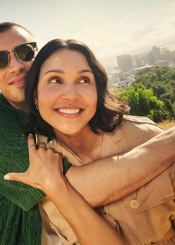Ein sich umarmendes, junges Paar auf einem Hügel vor der Skyline von Kapstadt.