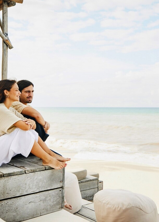 Ein Paar sitzt entspannt auf einer Holztribüne mit Polstermöbeln an einem weißen Sandstrand und blickt aufs Meer.
